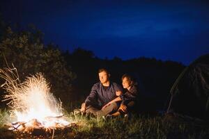 glücklich romantisch Paar Reisende ruhen beim Lagerfeuer in der Nähe von glühend Tourist Zelt unter tolle Nacht Himmel. das Konzept von aktiv Erholung und Reise mit ein Zelt foto