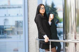 Porträt von erfolgreich Geschäft Frau gehen zu Arbeit mit Kaffee Gehen in der Nähe von Büro Gebäude auf Stadt Straße. foto