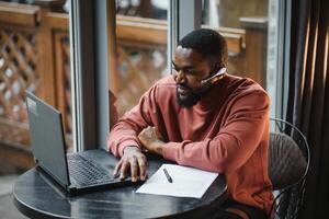 heiter jung afrikanisch amerikanisch Schüler Mann im Kopfhörer mit Computer im Cafe. online Video Plaudern mit Freunde. foto