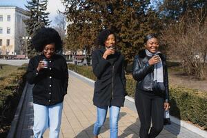 drei schön und stilvoll dunkelhäutig Mädchen mit lange Haar Stehen im ein Stadt und Trinken ein Kaffee und verwenden das Telefone foto