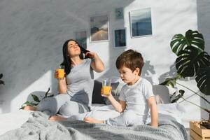 Mama mit Sohn im Pyjama Trinken Saft im das Morgen im Bett. gesund Essen Konzept foto