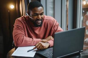 heiter jung afrikanisch amerikanisch Schüler Mann im Kopfhörer mit Computer im Cafe. online Video Plaudern mit Freunde. foto