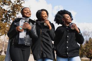 drei schön und stilvoll dunkelhäutig Mädchen mit lange Haar Stehen im ein Stadt und Trinken ein Kaffee und verwenden das Telefone foto