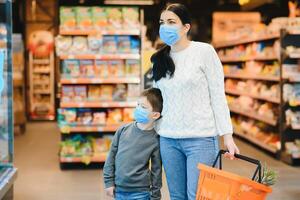 Einkaufen mit Kinder während Virus Ausbruch. Mutter und Kind tragen chirurgisch Gesicht Maske Kauf Obst im Supermarkt. Mama und wenig Junge Kaufen frisch Gemüse im Lebensmittelgeschäft speichern. Familie im Geschäft foto