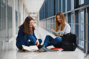 zwei ziemlich weiblich Studenten mit Bücher Sitzung auf das Fußboden im das Universität Flur foto