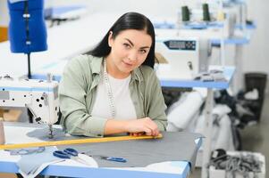 jung Schneider Frau näht Kleider auf Arbeiten Tisch. lächelnd Näherin und ihr Hand schließen oben im Werkstatt. foto