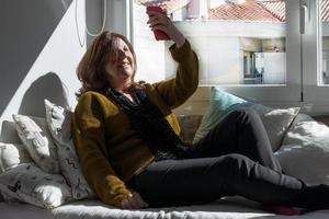 Kaukasische Frau im Alter von 50, die während eines Video-Chats mit einem Telefon lächelt. gelber Pullover und schwarze Hose. auf einem Sofa in der Nähe des Fensters. foto