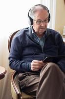 gealterter kaukasischer Mann zu Hause mit Brille, Kopfhörern und Blick auf sein Tablet foto