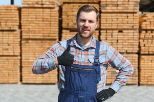 Porträt von ein gut aussehend Arbeiter wählen das Beste hölzern Bretter. Zimmermann Stehen Nächster zu ein groß Stapel von Holz Riegel im ein Lagerhaus. foto