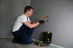 Elektriker Baumeister beim arbeiten, Installation von Steckdosen und Schalter. Fachmann im Overall mit ein Elektriker Werkzeug. gegen das Hintergrund von das Reparatur Seite? ˅. foto