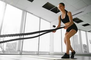 sportlich weiblich im ein Fitnessstudio Übungen mit Schlacht Seile während ihr Fitness trainieren Hohe Intensität Intervall Ausbildung. sie ist muskulös und verschwitzt foto