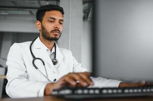 Menschen, Besetzung und Medizin Konzept. lächelnd männlich indisch arabisch Arzt im Weiß Mantel, Sitzung im medizinisch Büro beim Schreibtisch mit Laptop foto
