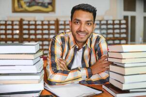 Porträt von heiter männlich International indisch Schüler mit Rucksack, Lernen Zubehör Stehen in der Nähe von Bücherregale beim Universität Bibliothek oder Buch Geschäft während brechen zwischen Unterricht. Bildung Konzept foto