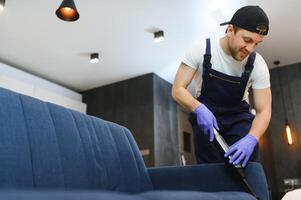 Reinigung Service. Mann Hausmeister im Handschuhe und Uniform Vakuum sauber Sofa mit Fachmann Ausrüstung. foto