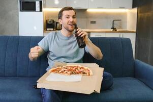 glücklich jung Mann Trinken Bier und Essen Pizza wann Aufpassen Spiel auf Fernseher beim Zuhause foto