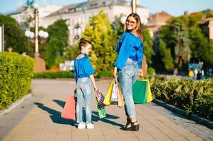 jung Mutter und ihr Tochter tun Einkaufen zusammen. Frau mit Mädchen Kind nach Einkaufen im Straße. Frau mit Tochter mit Einkaufen Taschen draußen. Frau und ihr Tochter nach Einkaufen foto