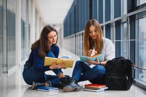 zwei ziemlich weiblich Studenten mit Bücher Sitzung auf das Fußboden im das Universität Flur foto