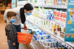 Einkaufen mit Kind während Virus Ausbruch. Mutter und Kind tragen chirurgisch Gesicht Maske Kauf Obst im Supermarkt. Mama und wenig Junge Kaufen frisch Gemüse im Lebensmittelgeschäft speichern. Familie im Geschäft foto