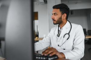 Menschen, Besetzung und Medizin Konzept. lächelnd männlich indisch arabisch Arzt im Weiß Mantel, Sitzung im medizinisch Büro beim Schreibtisch mit Laptop foto