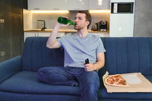 glücklich jung Mann Trinken Bier und Essen Pizza wann Aufpassen Spiel auf Fernseher beim Zuhause foto