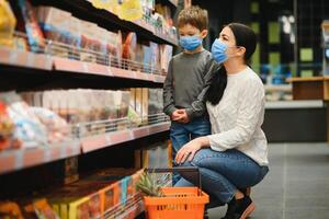 Mutter und ihr Sohn tragen schützend Gesicht Maske Geschäft beim ein Supermarkt während das Coronavirus Epidemie. foto