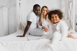 Porträt von glücklich gemischtrassig jung Familie Lügen auf gemütlich Weiß Bett beim heim, lächelnd International Mama und Papa entspannend mit wenig biracial Mädchen Kind posieren zum Bild im Schlafzimmer foto