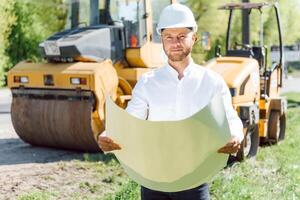 lächelnd Ingenieur mit Helm Stehen im Vorderseite von Bagger auf Straße Konstruktion Seite? ˅ foto