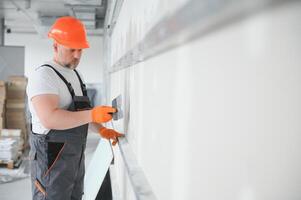 Mann Trockenbau Arbeiter oder Stuckateur Putten Gittergewebe Band zum Gipskarton auf ein Mauer mit ein Spatel und Gips. tragen Weiß Bauarbeiterhelm, Arbeit Handschuhe und Sicherheit Brille. Bild mit Kopieren Raum. foto