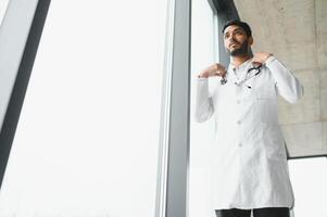 Porträt von männlich indisch Arzt tragen Weiß Mantel haben öffnen Tür auf Klinik Gang wie Hintergrund foto