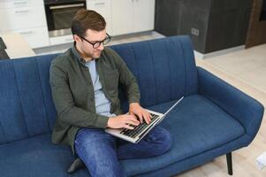 Männer Arbeiten auf Laptop Computer im seine Zimmer. Zuhause Arbeit oder lernen, freiberuflich Konzept. jung Mann Sitzung entspannt auf Sofa mit Laptop. modern Geschäftsmann mit Laptop. foto