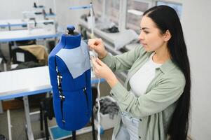 jung Schneider Frau näht Kleider auf Arbeiten Tisch. lächelnd Näherin und ihr Hand schließen oben im Werkstatt. foto