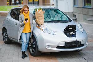 stilvoll Frau nach Kauf Produkte mit ein Einkaufen Tasche ist Stehen in der Nähe von das Laden elektrisch Wagen. foto