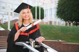 glücklich süß kaukasisch grad Mädchen ist lächelnd. sie ist im ein schwarz Granatwerfer Planke, mit rot Quaste, im Kleid, mit nett braun lockig Haar, Diplom im Hand foto