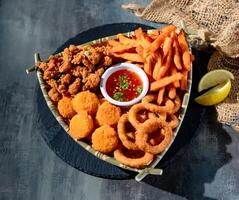 Snacker Teller mit Zwiebel Ring, Französisch Fritten, golden Jakobsmuscheln, Chili Soße im ein Gericht oben Aussicht auf dunkel Hintergrund foto