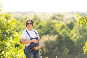 Porträt von ein glücklich und stolz schwanger Frau suchen beim ihr Bauch im ein Park beim Sonnenaufgang mit ein warm zurück Licht im das Hintergrund foto
