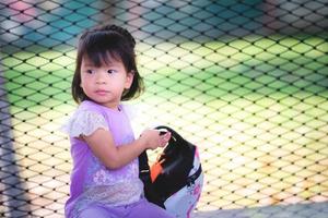 asiatisches entzückendes Kindermädchen sitzt auf einem Marmorsitz. eine Tasche halten und etwas anschauen. sie trug ein lila Kleid, weiße Spitzenärmel und blaue Schuhe. der Hintergrund ist ein Rasen und eine Balustrade. foto