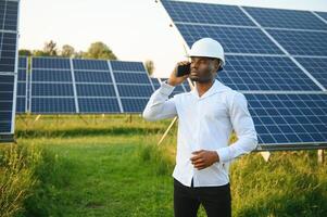 ein Afroamerikaner Ingenieur im ein Weiß Hemd und schwer Hut ist Arbeiten auf ein Feld von Solar- Tafeln. Solar- verlängerbar Energie Konzept. foto