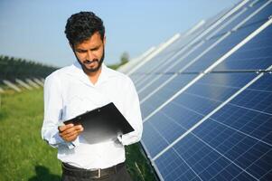 Porträt von jung indisch männlich Ingenieur Stehen in der Nähe von Solar- Tafeln, mit klar Blau Himmel Hintergrund, verlängerbar und sauber Energie. Fertigkeit Indien, Kopieren Raum. foto