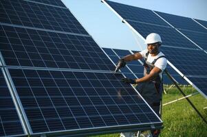 Afroamerikaner Arbeiter im grau Overall und ein Weiß schwer Hut funktioniert im ein Feld von Solar- Tafeln. Solar- verlängerbar Energie Konzept. foto