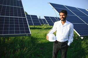 Porträt von jung indisch männlich Ingenieur Stehen in der Nähe von Solar- Tafeln, mit klar Blau Himmel Hintergrund, verlängerbar und sauber Energie. Fertigkeit Indien, Kopieren Raum. foto