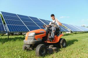 ein Mann Arbeiten beim Solar- Leistung Bahnhof. ein Arbeiter auf ein Garten Traktor mäht Gras auf ein Solar- Panel Bauernhof. foto