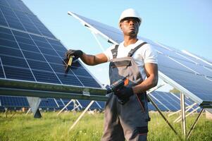 Afroamerikaner Arbeiter im grau Overall und ein Weiß schwer Hut funktioniert im ein Feld von Solar- Tafeln. Solar- verlängerbar Energie Konzept. foto