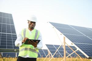 ein Afroamerikaner Ingenieur im ein Weiß Hemd und schwer Hut ist Arbeiten auf ein Feld von Solar- Tafeln. Solar- verlängerbar Energie Konzept. foto