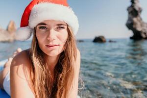 Frau Meer sup. schließen oben Porträt von glücklich jung kaukasisch Frau mit lange Haar im Santa Hut suchen beim Kamera und lächelnd. süß Frau Porträt im ein Weiß Bikini posieren auf sup Tafel im das Meer foto