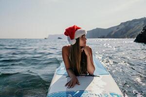 Frau Meer sup. schließen oben Porträt von glücklich jung kaukasisch Frau mit lange Haar im Santa Hut suchen beim Kamera und lächelnd. süß Frau Porträt im ein Weiß Bikini posieren auf sup Tafel im das Meer foto