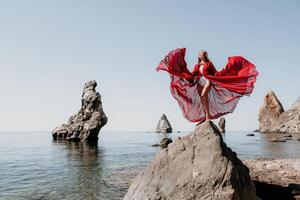 Frau Reise Meer. jung glücklich Frau im ein lange rot Kleid posieren auf ein Strand in der Nähe von das Meer auf Hintergrund von vulkanisch Felsen, mögen im Island, Teilen Reise Abenteuer Reise foto