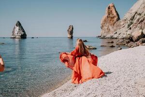 Frau Reise Meer. glücklich Tourist im rot Kleid genießen nehmen Bild draußen zum Erinnerungen. Frau Reisender posieren auf das Felsen beim Meer Bucht umgeben durch vulkanisch Berge, Teilen Reise Abenteuer Reise foto