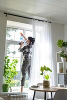 Frau manuell wäscht das Fenster von das Haus mit ein Lappen mit sprühen Reiniger und Mopp Innerhalb das Innere mit Weiß Vorhänge. Wiederherstellen bestellen und Sauberkeit im das Frühling, Reinigung warten foto