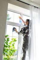 Frau manuell wäscht das Fenster von das Haus mit ein Lappen mit sprühen Reiniger und Mopp Innerhalb das Innere mit Weiß Vorhänge. Wiederherstellen bestellen und Sauberkeit im das Frühling, Reinigung warten foto