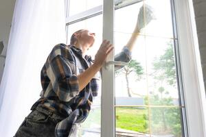 Frau manuell wäscht das Fenster von das Haus mit ein Lappen mit sprühen Reiniger und Mopp Innerhalb das Innere mit Weiß Vorhänge. Wiederherstellen bestellen und Sauberkeit im das Frühling, Reinigung warten foto